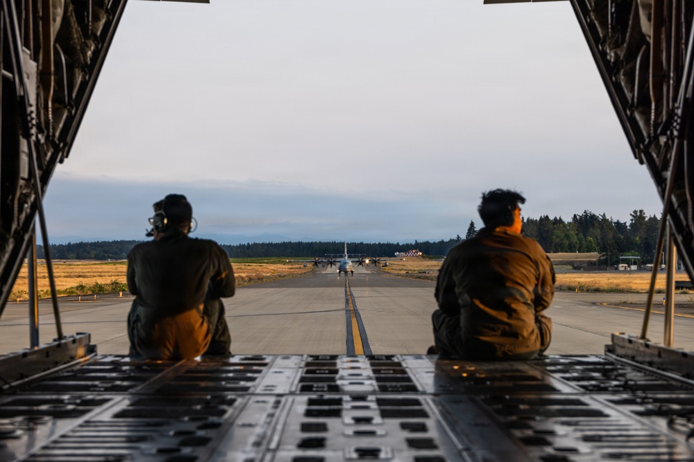 Two military personnel are seated on the open rear cargo ramp of a large military transport aircraft, looking out onto a runway. In the distance, another military turboprop aircraft — consistent with a C-130 Hercules — is visible on the taxiway or runway. The scene appears to be at a military airfield surrounded by forested terrain, photographed during golden hour lighting.