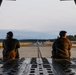 Two military personnel are seated on the open rear cargo ramp of a large military transport aircraft, looking out onto a runway. In the distance, another military turboprop aircraft — consistent with a C-130 Hercules — is visible on the taxiway or runway. The scene appears to be at a military airfield surrounded by forested terrain, photographed during golden hour lighting.