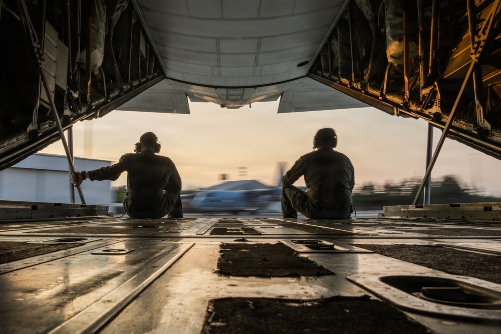 Two individuals are seated on the open rear cargo ramp of a large military transport aircraft, viewed from inside looking outward toward a tarmac or airfield at dusk or dawn. The silhouetted figures face away from the camera, observing the exterior environment with a motion-blurred vehicle visible in the background. The warm ambient light and industrial interior of the cargo bay create a dramatic compositional contrast.