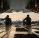 Two individuals are seated on the open rear cargo ramp of a large military transport aircraft, viewed from inside looking outward toward a tarmac or airfield at dusk or dawn. The silhouetted figures face away from the camera, observing the exterior environment with a motion-blurred vehicle visible in the background. The warm ambient light and industrial interior of the cargo bay create a dramatic compositional contrast.