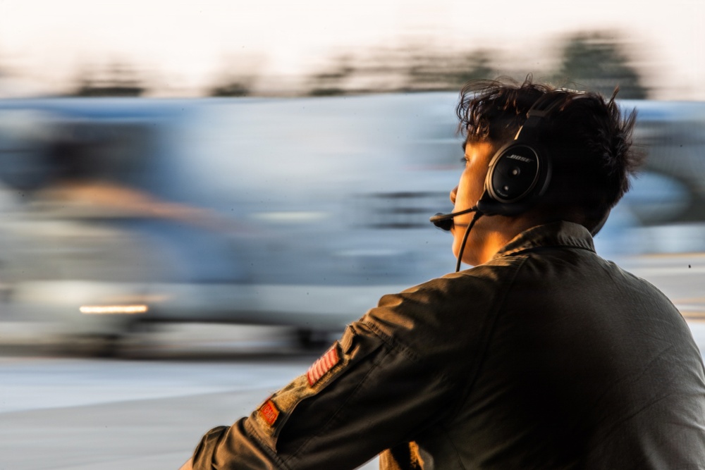 A person wearing a Bose aviation headset and an olive/green flight suit with a US flag patch on the sleeve is observed from behind and slightly to the side. They appear to be on a flight line or airfield, watching a fast-moving aircraft that is heavily motion-blurred in the background. The image uses a slow shutter speed or panning technique to convey the speed of the aircraft.