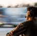 A person wearing a Bose aviation headset and an olive/green flight suit with a US flag patch on the sleeve is observed from behind and slightly to the side. They appear to be on a flight line or airfield, watching a fast-moving aircraft that is heavily motion-blurred in the background. The image uses a slow shutter speed or panning technique to convey the speed of the aircraft.