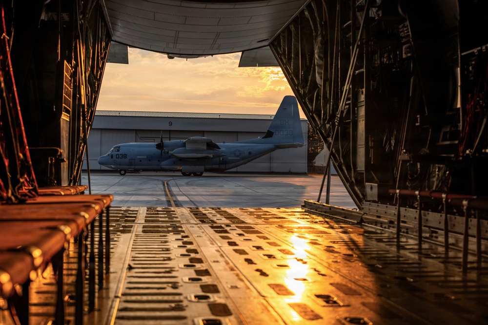 The image is taken from inside the open cargo ramp of a large military transport aircraft, looking out onto a flight line at sunset or sunrise. A C-130 Hercules turboprop transport aircraft bearing the number '038' is visible parked on the tarmac outside, with a hangar marked '9' in the background. The warm golden light reflects off the wet cargo ramp floor, creating a dramatic and atmospheric composition.