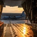 The image is taken from inside the open cargo ramp of a large military transport aircraft, looking out onto a flight line at sunset or sunrise. A C-130 Hercules turboprop transport aircraft bearing the number '038' is visible parked on the tarmac outside, with a hangar marked '9' in the background. The warm golden light reflects off the wet cargo ramp floor, creating a dramatic and atmospheric composition.
