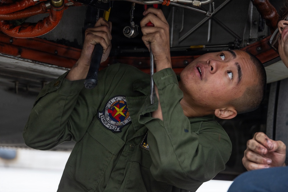 A military aviation technician in an olive green flight suit is performing maintenance work on the underside of an aircraft. The individual is using hand tools, including what appears to be a ratchet or torque wrench, while looking upward at the mechanical components. A unit patch reading 'VMGR-152' is visible on the left shoulder of the flight suit.