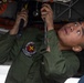 A military aviation technician in an olive green flight suit is performing maintenance work on the underside of an aircraft. The individual is using hand tools, including what appears to be a ratchet or torque wrench, while looking upward at the mechanical components. A unit patch reading 'VMGR-152' is visible on the left shoulder of the flight suit.