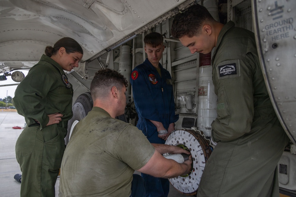 Four military personnel are performing maintenance on an aircraft, likely a military patrol or transport aircraft. One technician in olive/green flight suits is kneeling and handling a circular mechanical component, possibly a wheel brake assembly or rotor disc, while three others observe. The work is taking place in an open wheel well or equipment bay area on a flight line.