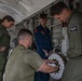 Four military personnel are performing maintenance on an aircraft, likely a military patrol or transport aircraft. One technician in olive/green flight suits is kneeling and handling a circular mechanical component, possibly a wheel brake assembly or rotor disc, while three others observe. The work is taking place in an open wheel well or equipment bay area on a flight line.
