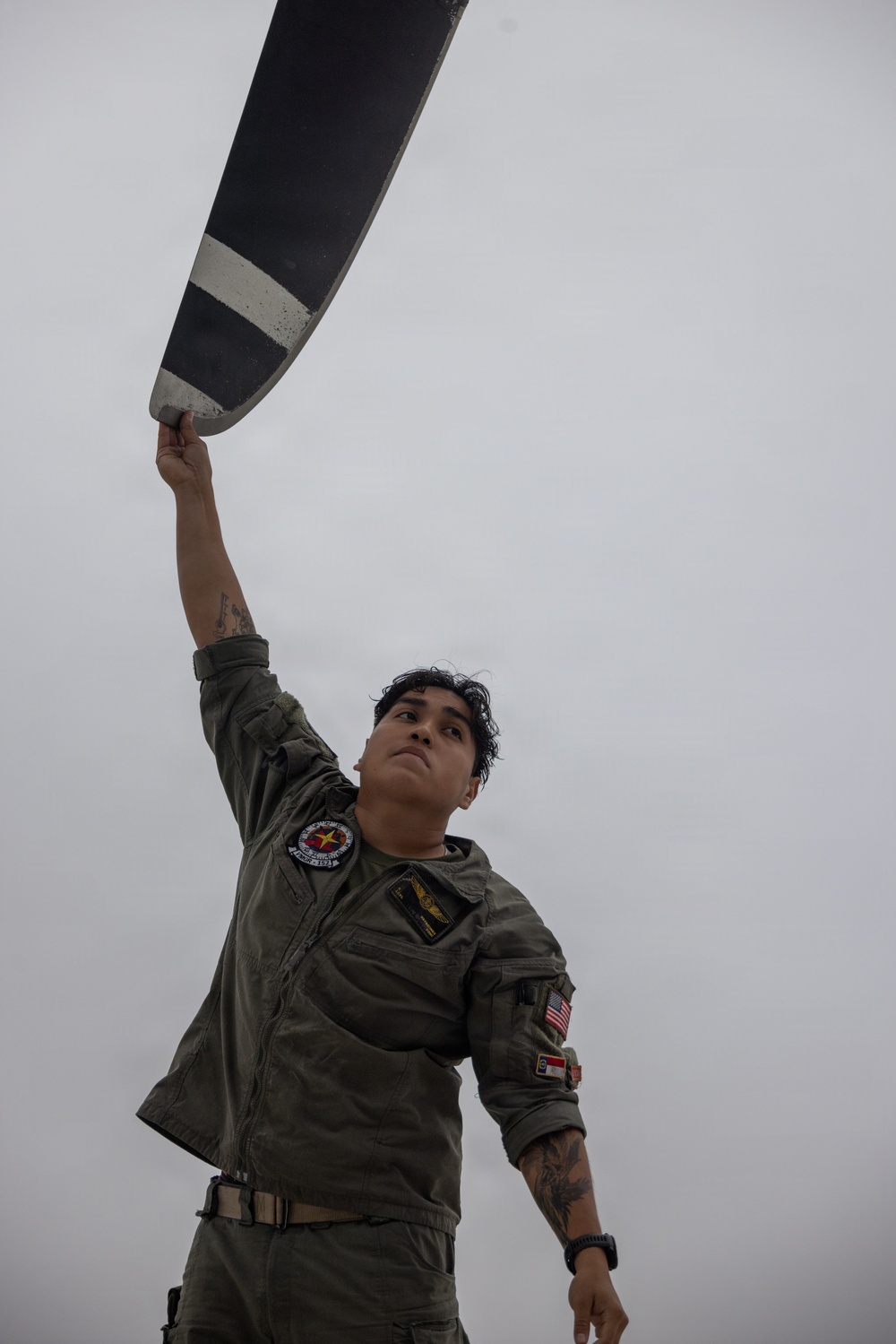 A military aviator in an olive drab flight suit is photographed from a low angle while gripping what appears to be a propeller blade or aircraft control surface with one raised arm. The individual wears multiple patches including a US flag patch, a state flag patch (appears to be North Carolina), a unit patch, and aviator wings on the chest. The dramatic upward angle and overcast sky create a striking compositional effect.