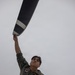 A military aviator in an olive drab flight suit is photographed from a low angle while gripping what appears to be a propeller blade or aircraft control surface with one raised arm. The individual wears multiple patches including a US flag patch, a state flag patch (appears to be North Carolina), a unit patch, and aviator wings on the chest. The dramatic upward angle and overcast sky create a striking compositional effect.
