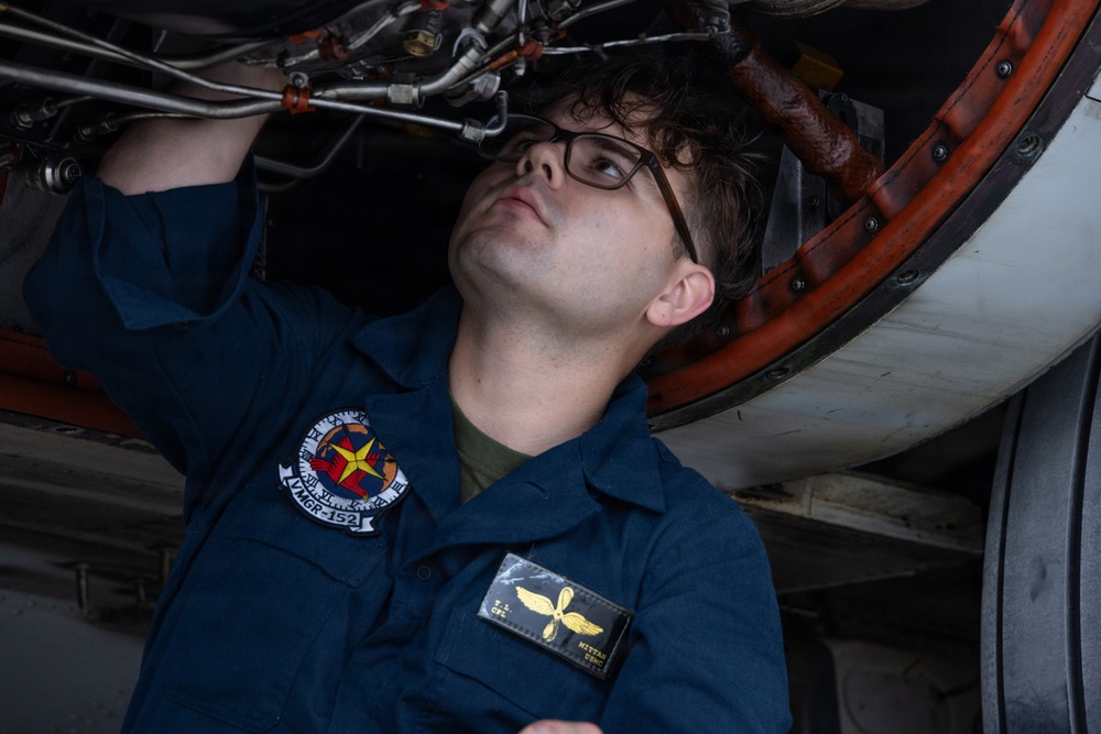 A young military aviation mechanic is performing maintenance work underneath an aircraft engine or landing gear assembly. The individual is wearing a Navy/Marine Corps coverall uniform with a unit patch and name tag, working in what appears to be a hangar environment. They are looking upward while using both hands to inspect or adjust components.
