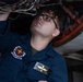 A young military aviation mechanic is performing maintenance work underneath an aircraft engine or landing gear assembly. The individual is wearing a Navy/Marine Corps coverall uniform with a unit patch and name tag, working in what appears to be a hangar environment. They are looking upward while using both hands to inspect or adjust components.