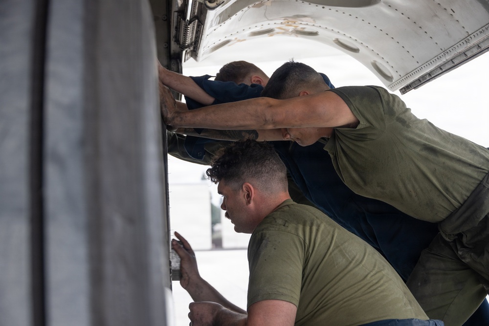 Three individuals are performing maintenance or inspection work on what appears to be a military or commercial aircraft. They are leaning into an open panel or access bay on the underside or side of the aircraft fuselage. The workers are wearing olive green and dark blue t-shirts, suggesting a military maintenance crew.