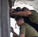 Three individuals are performing maintenance or inspection work on what appears to be a military or commercial aircraft. They are leaning into an open panel or access bay on the underside or side of the aircraft fuselage. The workers are wearing olive green and dark blue t-shirts, suggesting a military maintenance crew.