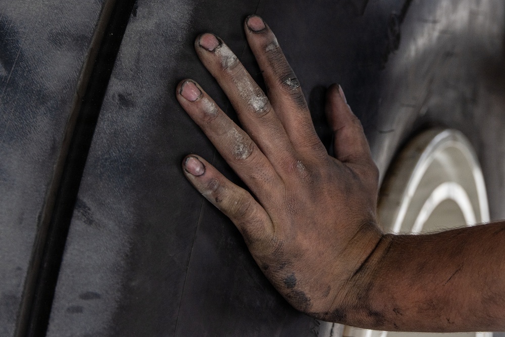 A close-up photograph of a worker's hand pressed against a dark vehicle body panel near a wheel well. The hand is visibly dirty with grime, dust, and residue on the fingers and knuckles, indicating manual labor. A silver alloy wheel is partially visible in the background to the right.