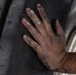 A close-up photograph of a worker's hand pressed against a dark vehicle body panel near a wheel well. The hand is visibly dirty with grime, dust, and residue on the fingers and knuckles, indicating manual labor. A silver alloy wheel is partially visible in the background to the right.
