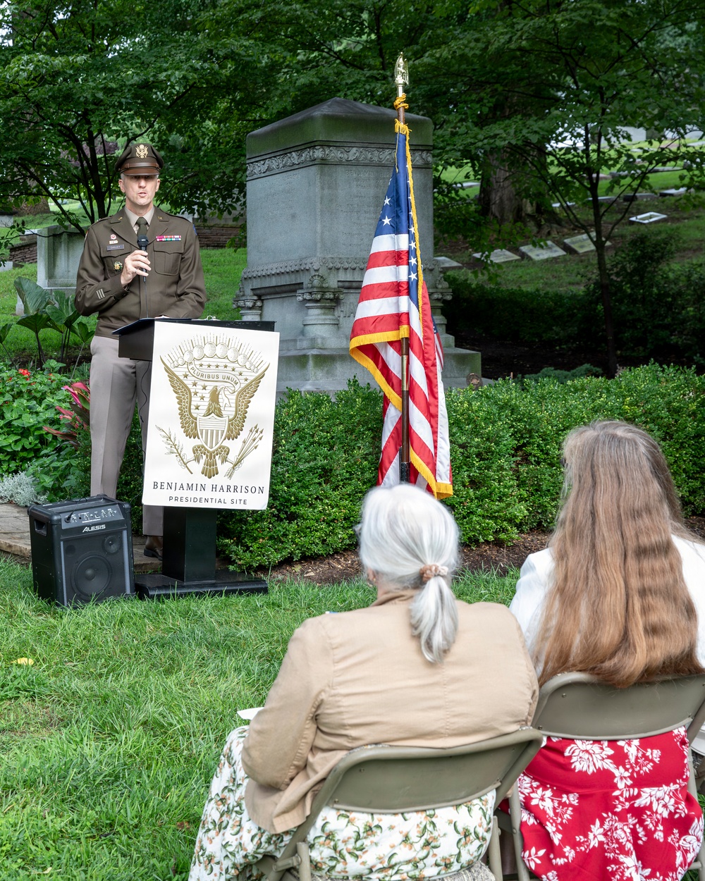 An outdoor ceremony is taking place at the Benjamin Harrison Presidential Site, likely at a cemetery or memorial grounds. A U.S. Army officer in dress uniform is speaking at a podium bearing the Benjamin Harrison Presidential Site seal, with an American flag displayed beside him. Two attendees are seated on folding chairs in the foreground, listening to the address.