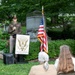 An outdoor ceremony is taking place at the Benjamin Harrison Presidential Site, likely at a cemetery or memorial grounds. A U.S. Army officer in dress uniform is speaking at a podium bearing the Benjamin Harrison Presidential Site seal, with an American flag displayed beside him. Two attendees are seated on folding chairs in the foreground, listening to the address.