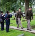 A military memorial ceremony is taking place in a historic cemetery with green grass and old grave markers. Personnel in modern U.S. Army service uniforms are walking along a stone pathway while being saluted by individuals dressed in Civil War-era Union Army uniforms. The scene suggests a commemorative or wreath-laying event honoring fallen soldiers.