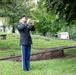 A U.S. Army soldier in dress uniform is playing a bugle or trumpet in a cemetery, likely performing 'Taps' during a military funeral or memorial ceremony. The soldier stands on a grassy area surrounded by grave markers, monuments, and mature trees. The setting and formal attire indicate this is an official military honors ceremony.