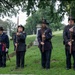 Four individuals dressed in Union Army Civil War-era reenactment uniforms stand at attention in a cemetery setting. Three are holding period-accurate musket rifles at port arms or order arms positions, while the leftmost figure holds a sword. The setting, costumes, and formal posture suggest this is a memorial or commemorative ceremony.