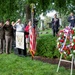 A formal memorial ceremony is taking place at what appears to be the Benjamin Harrison Presidential Site, identifiable by the podium insignia. Attendees including military personnel in uniform, civilians, and officials are gathered in a cemetery setting, with some saluting and others placing hands over hearts. A patriotic wreath adorned with red, white, and blue flowers and ribbons stands prominently on a stand to the right.