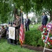 A formal memorial ceremony is taking place at the Benjamin Harrison Presidential Site, likely a cemetery or burial ground. A military officer in Army dress uniform is delivering remarks at a podium bearing the Benjamin Harrison Presidential Site seal, flanked by an American flag. Dignitaries, military personnel, and civilians are seated and standing in attendance, with a patriotic red, white, and blue floral wreath displayed prominently.