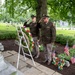 Two military personnel in olive drab dress uniforms are rendering a salute at a cemetery memorial ceremony. They are standing before a red, white, and blue wreath mounted on a white easel, surrounded by small American flags and floral arrangements. The setting appears to be a well-maintained civilian cemetery with historic grave markers and mature trees in the background.