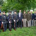 A group of approximately ten men pose for a formal photograph in what appears to be a cemetery setting. The group includes individuals in Civil War-era Union Army reenactment uniforms carrying period rifles, men in modern U.S. Army dress uniforms with medals, civilians in suits and blazers with service ribbons, and one man in a business suit and tie. The setting and attire suggest a military memorial or commemorative ceremony.