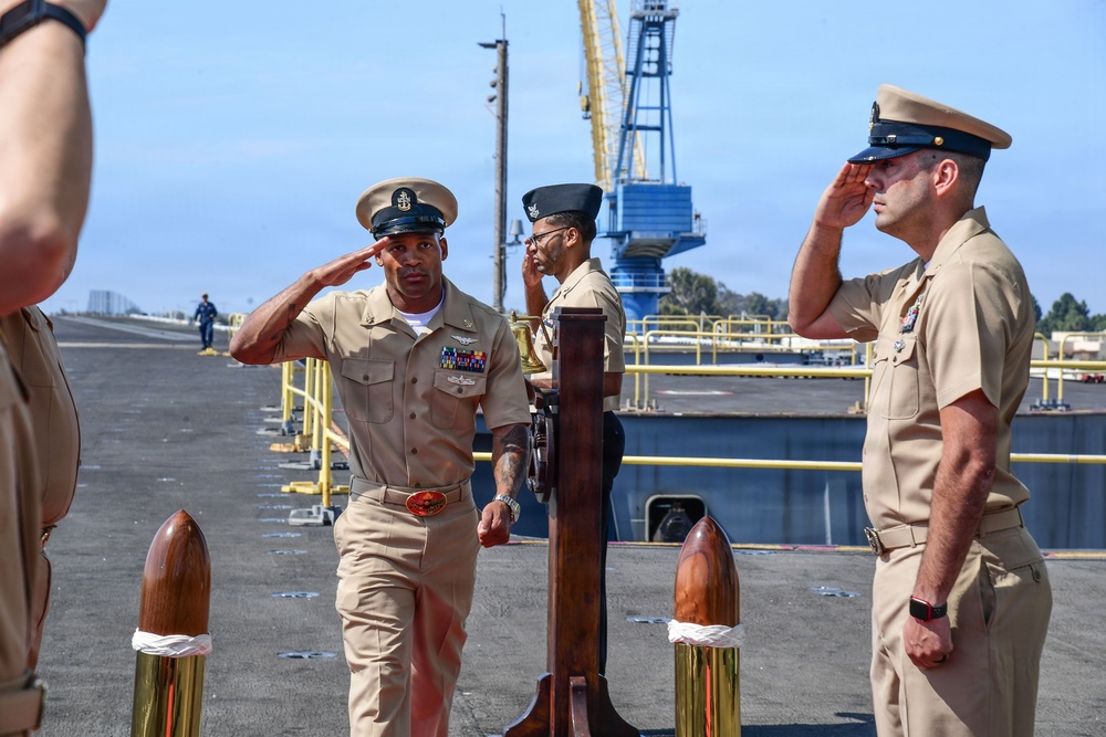 A formal U.S. Navy ceremony is taking place on what appears to be a ship's deck or pier, with personnel in Service Khaki uniforms exchanging salutes. Two senior enlisted or officer personnel face each other in a saluting exchange near a ship's bell mounted on a wooden stand, suggesting a change of command or boarding ceremony. The setting includes a naval installation with a large blue crane visible in the background.