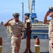 A formal U.S. Navy ceremony is taking place on what appears to be a ship's deck or pier, with personnel in Service Khaki uniforms exchanging salutes. Two senior enlisted or officer personnel face each other in a saluting exchange near a ship's bell mounted on a wooden stand, suggesting a change of command or boarding ceremony. The setting includes a naval installation with a large blue crane visible in the background.
