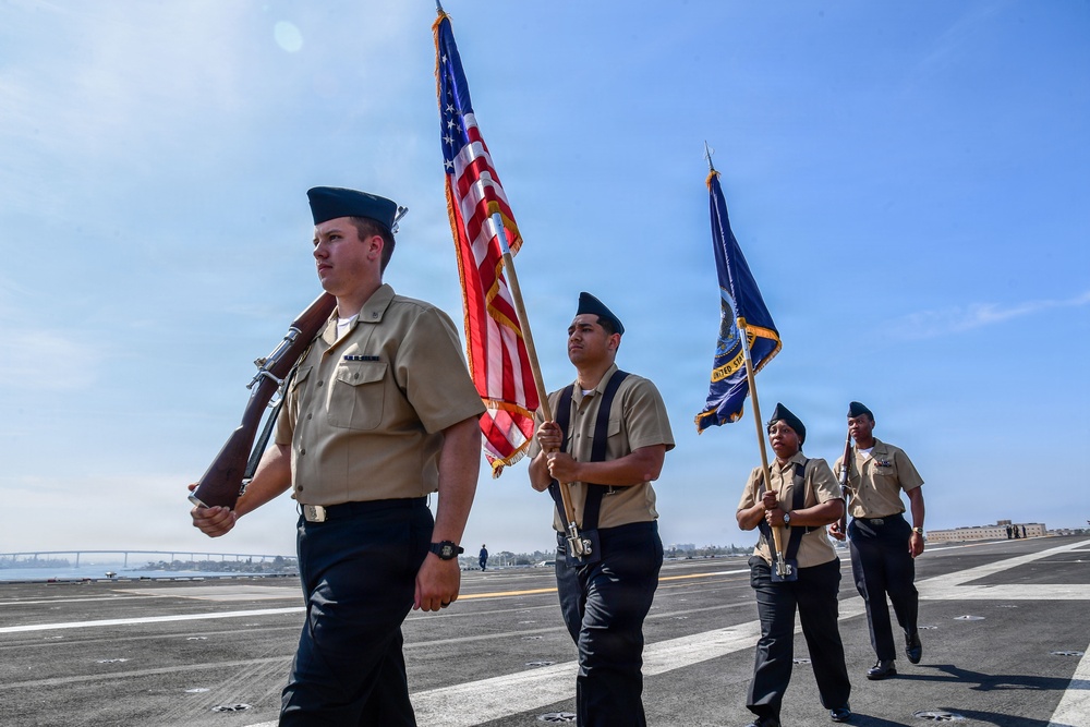 A U.S. Navy color guard is marching in formation on what appears to be the flight deck of an aircraft carrier. The personnel are in service khaki uniforms with garrison caps, carrying the American flag, a Navy unit flag, and a ceremonial rifle. A bridge and city skyline are visible in the background, suggesting a port location likely in San Diego, California.