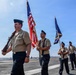 A U.S. Navy color guard is marching in formation on what appears to be the flight deck of an aircraft carrier. The personnel are in service khaki uniforms with garrison caps, carrying the American flag, a Navy unit flag, and a ceremonial rifle. A bridge and city skyline are visible in the background, suggesting a port location likely in San Diego, California.