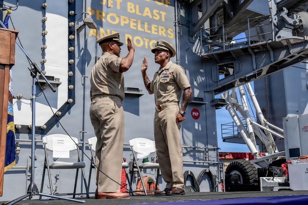 Two U.S. Navy personnel in khaki service uniforms are conducting a military ceremony, likely a swearing-in or re-enlistment, aboard a naval vessel. Both individuals have their right hands raised in an oath-taking gesture on a stage. Reserved chairs labeled 'GUNNER KITTLES' and 'PRESIDING OFFICER' are visible in the background.