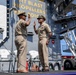 Two U.S. Navy personnel in khaki service uniforms are conducting a military ceremony, likely a swearing-in or re-enlistment, aboard a naval vessel. Both individuals have their right hands raised in an oath-taking gesture on a stage. Reserved chairs labeled 'GUNNER KITTLES' and 'PRESIDING OFFICER' are visible in the background.