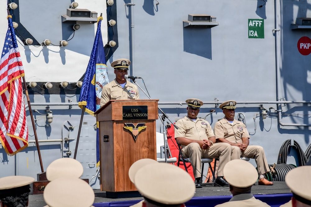 A formal U.S. Navy ceremony is taking place on the deck of the USS Carl Vinson, an aircraft carrier. A naval officer in service khaki uniform is speaking at a podium bearing the ship's name and insignia, while two other officers are seated behind him. An audience of sailors in service dress white covers is visible in the foreground.