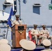 A formal U.S. Navy ceremony is taking place on the deck of the USS Carl Vinson, an aircraft carrier. A naval officer in service khaki uniform is speaking at a podium bearing the ship's name and insignia, while two other officers are seated behind him. An audience of sailors in service dress white covers is visible in the foreground.