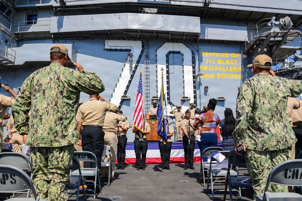 A formal military ceremony is taking place on the flight deck of a U.S. Navy aircraft carrier, hull number 70. Personnel in various uniforms are rendering salutes toward a podium flanked by the American flag and a Navy flag. Civilians, likely family members, are also present at the ceremony.