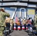 A formal military ceremony is taking place on the flight deck of a U.S. Navy aircraft carrier, hull number 70. Personnel in various uniforms are rendering salutes toward a podium flanked by the American flag and a Navy flag. Civilians, likely family members, are also present at the ceremony.