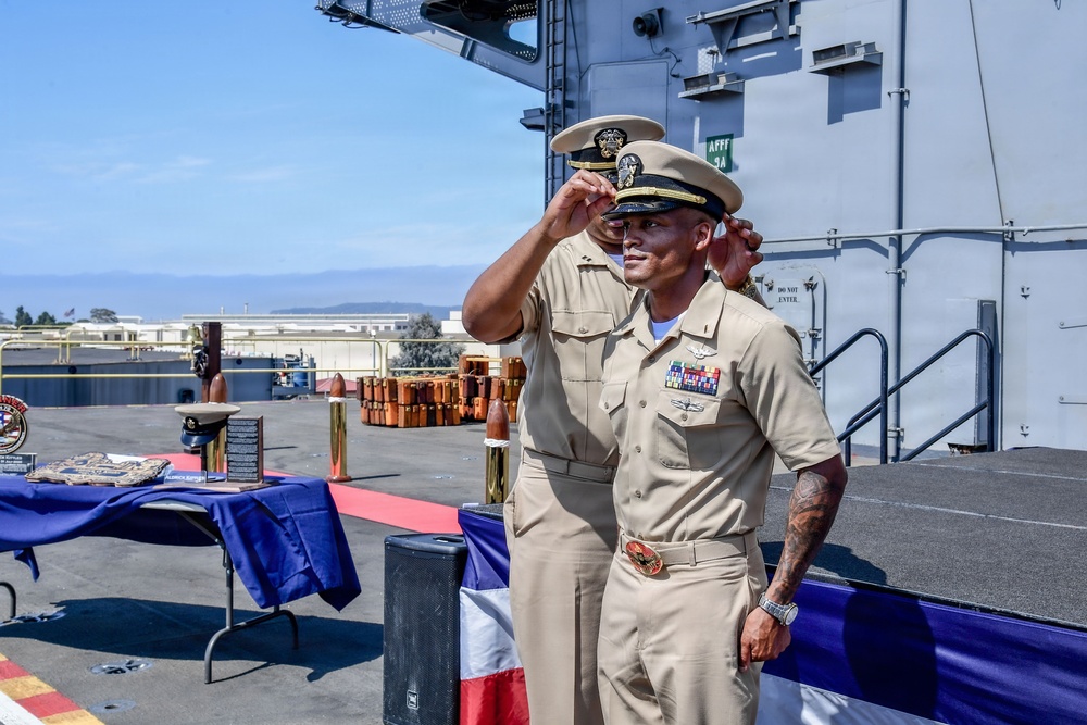 Two U.S. Navy personnel in khaki service uniforms are participating in what appears to be a formal military ceremony on the deck of a naval vessel. One individual is adjusting or placing a cover (hat) on the other, suggesting a promotion, retirement, or change of command ceremony. A ceremonial table with plaques, awards, and a blue tablecloth is visible in the background.