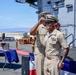 Two U.S. Navy personnel in khaki service uniforms are participating in what appears to be a formal military ceremony on the deck of a naval vessel. One individual is adjusting or placing a cover (hat) on the other, suggesting a promotion, retirement, or change of command ceremony. A ceremonial table with plaques, awards, and a blue tablecloth is visible in the background.