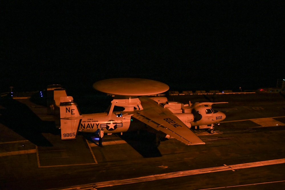 A nighttime photograph taken on the flight deck of USS Carl Vinson (CVN-70), showing two E-2C/D Hawkeye airborne early warning aircraft illuminated by artificial orange lighting. The foreground aircraft bears bureau number 9865 and is marked with 'NAVY' and 'USS CARL VINSON' text, while a second aircraft numbered 602 is visible to the right with wings folded. Both aircraft display the distinctive rotodome radar disc mounted above the fuselage.