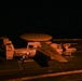 A nighttime photograph taken on the flight deck of USS Carl Vinson (CVN-70), showing two E-2C/D Hawkeye airborne early warning aircraft illuminated by artificial orange lighting. The foreground aircraft bears bureau number 9865 and is marked with 'NAVY' and 'USS CARL VINSON' text, while a second aircraft numbered 602 is visible to the right with wings folded. Both aircraft display the distinctive rotodome radar disc mounted above the fuselage.