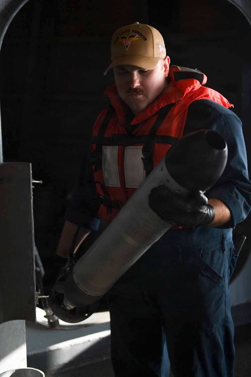 A U.S. Navy sailor assigned to USS Carl Vinson (CVN-70) is handling a cylindrical object, likely a flare canister or similar deck equipment. He is wearing a bright orange flotation/life vest over blue coveralls and black gloves, indicating work in a potentially hazardous deck environment. The setting appears to be aboard a naval vessel in a shadowed deck or hangar bay area.