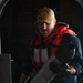 A U.S. Navy sailor assigned to USS Carl Vinson (CVN-70) is handling a cylindrical object, likely a flare canister or similar deck equipment. He is wearing a bright orange flotation/life vest over blue coveralls and black gloves, indicating work in a potentially hazardous deck environment. The setting appears to be aboard a naval vessel in a shadowed deck or hangar bay area.