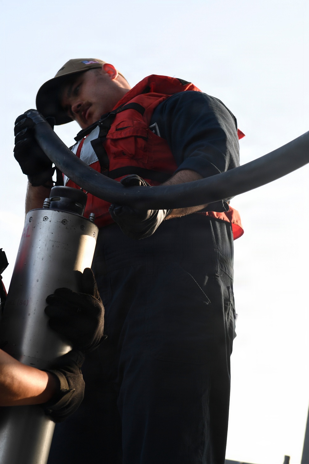 A person wearing a red inflatable life vest, dark coveralls, a cap, and black gloves is handling a large hose connected to a metal cylindrical fitting or nozzle. The low-angle shot looks up at the worker against a pale sky background. The scene appears to be a maritime or naval fueling/transfer operation.