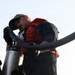 A person wearing a red inflatable life vest, dark coveralls, a cap, and black gloves is handling a large hose connected to a metal cylindrical fitting or nozzle. The low-angle shot looks up at the worker against a pale sky background. The scene appears to be a maritime or naval fueling/transfer operation.