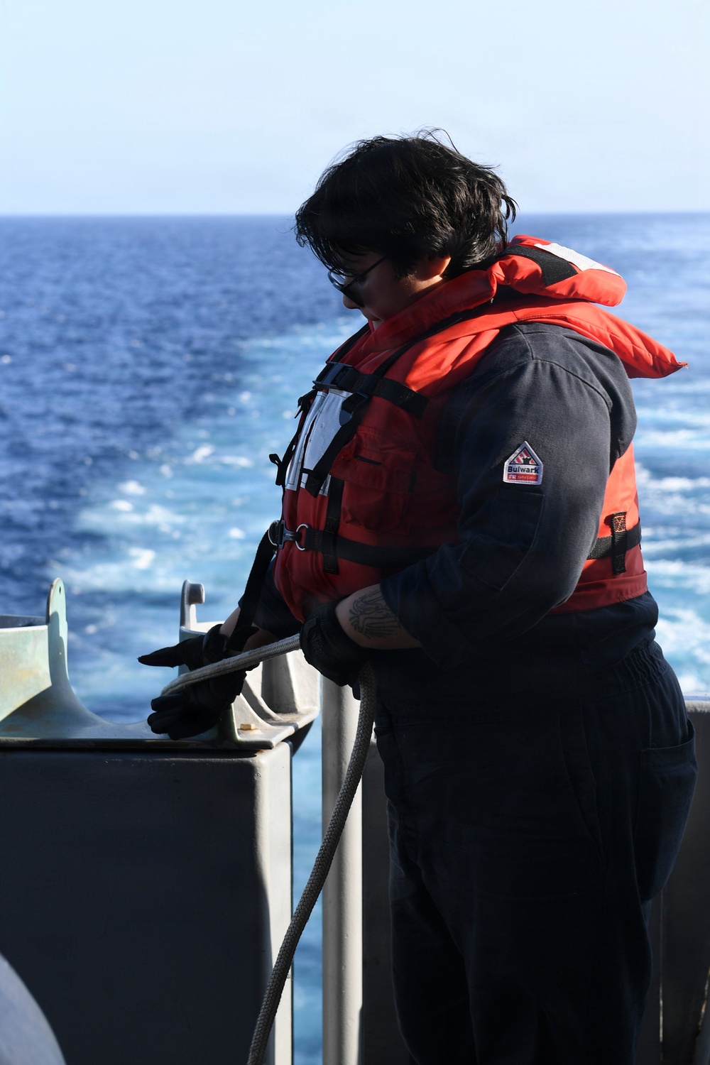 A person wearing a red life vest and dark navy coveralls stands on the deck of a vessel at sea, handling a rope or line near a metal railing. The open ocean is visible in the background with white wake indicating the vessel is underway. The individual is wearing black gloves and sunglasses and appears to be performing a deck seamanship task.