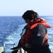 A person wearing a red life vest and dark navy coveralls stands on the deck of a vessel at sea, handling a rope or line near a metal railing. The open ocean is visible in the background with white wake indicating the vessel is underway. The individual is wearing black gloves and sunglasses and appears to be performing a deck seamanship task.