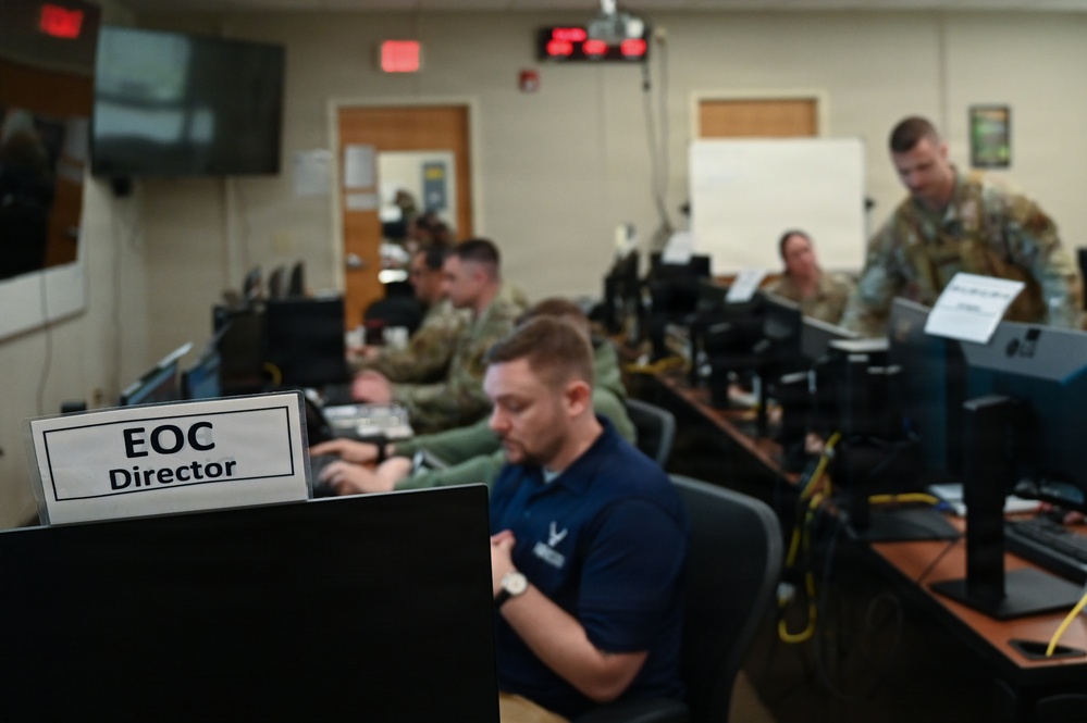 The image shows personnel working inside an Emergency Operations Center (EOC), as indicated by a nameplate reading 'EOC Director' in the foreground. Multiple individuals in military uniforms (camouflage) and civilian attire are seated at workstations with computers. The setting appears to be an active operations or exercise environment with monitoring equipment and displays visible.